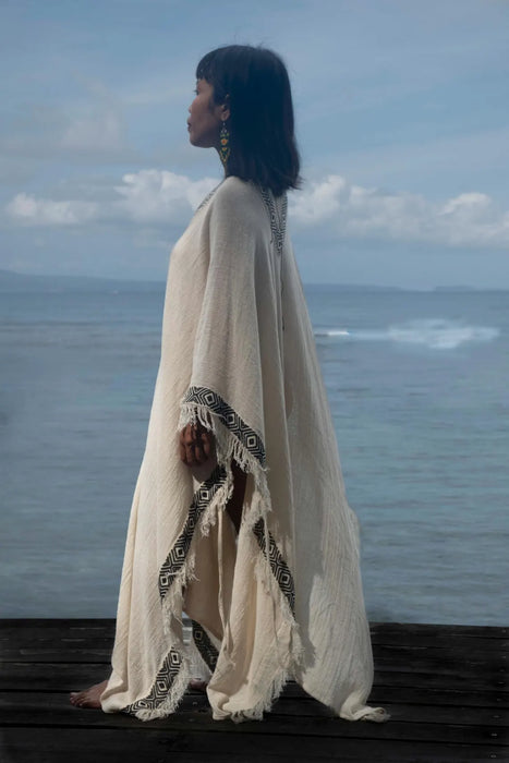Woman in a long, light-colored dress with a patterned border standing on a dock by the ocean.