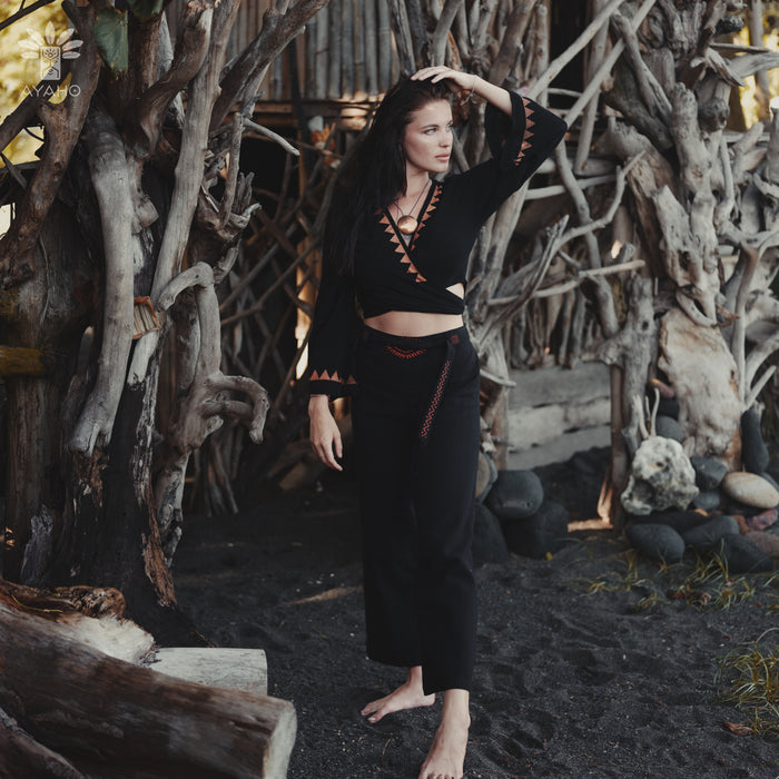 Woman in a black outfit standing in a natural setting with wooden structures and rocks.