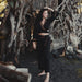 Woman in a black outfit standing in a natural setting with wooden structures and rocks.
