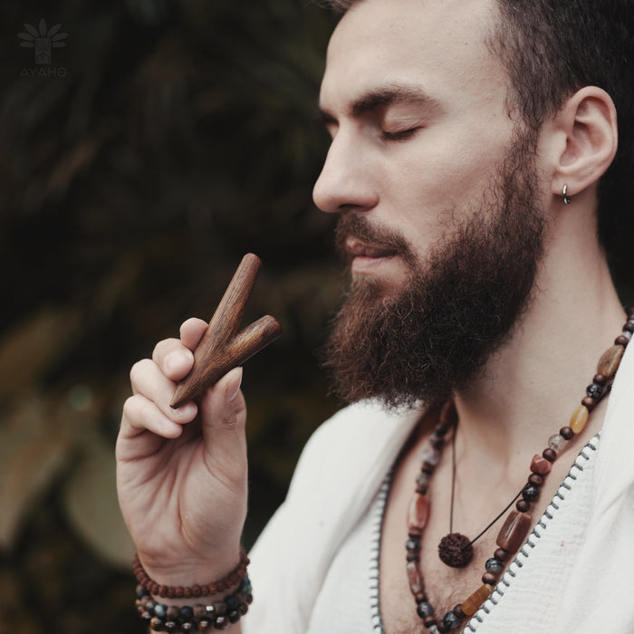 Man with a beard holding a wooden object, wearing a necklace and bracelets, with a blurred natural background.
