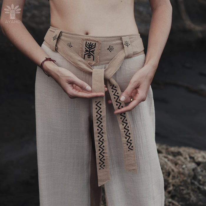 Person wearing a beige belt with black patterns, standing against a natural background.