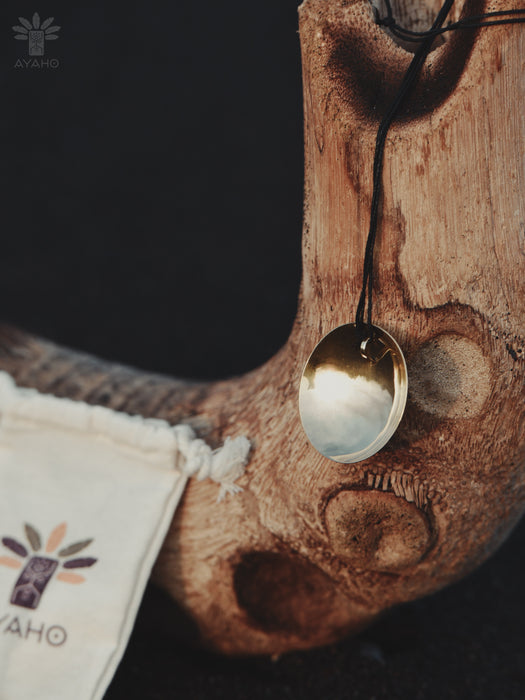 Gold pendant on a black cord hanging from a wooden surface with 'AYAHO' branding.