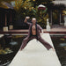 Man in traditional attire performing a martial arts pose by a pool with lush greenery in the background