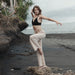 Woman in black top and beige pants standing on a rocky beach with ocean and sky in the background