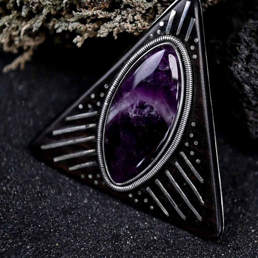 Striking silver triangular amulet with a richly hued amethyst center, designed for harmony, surrounded by symbolic engravings on rosewood for spiritual grounding.