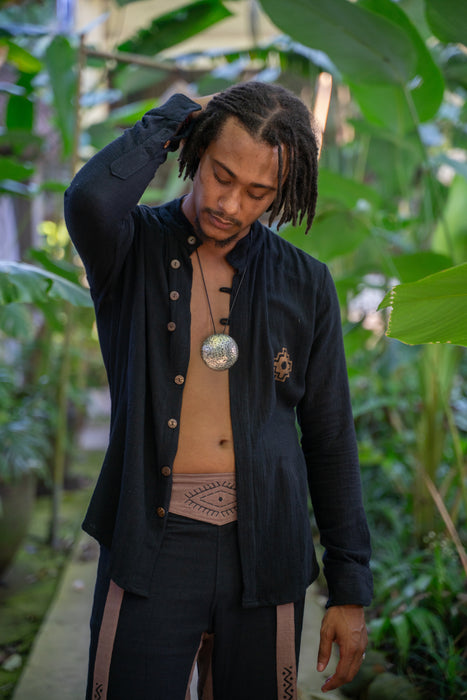 Man in a black jacket with buttons, beige shirt, and dark pants standing in a lush green outdoor setting.