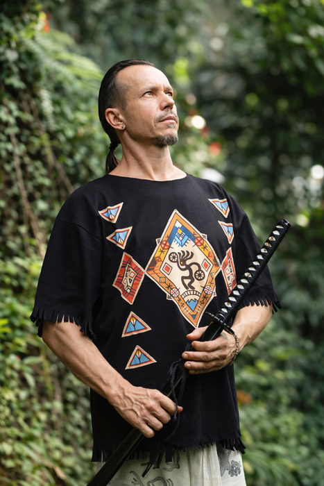 Man wearing a black embroidered shirt holding a weapon with a green, leafy background