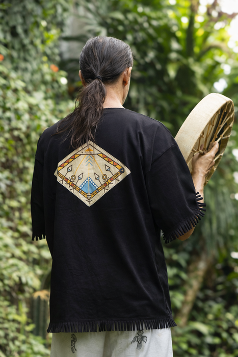 Person wearing a black shirt with a geometric design on the back, holding a mat in a forest setting.