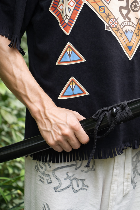 Person wearing a black garment with colorful geometric patterns, holding a black object against a blurred natural background.