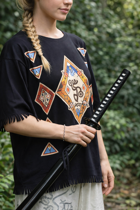 Person wearing a black poncho with geometric patterns holding a sword against a green leafy background