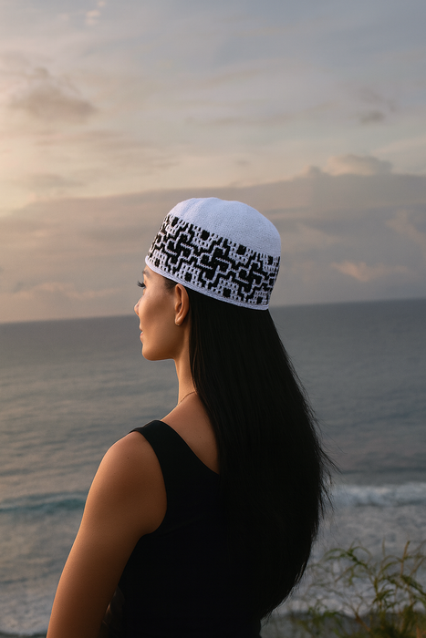 Woman wearing a white headscarf with black patterns, standing by the ocean at sunset.