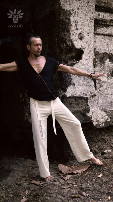 Man in black top and white pants standing with arms outstretched against a textured wall.