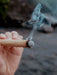 Hand holding a burning incense stick with smoke against a blurred natural background