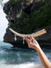 Hand holding a curved piece of driftwood with a small ornament against a rocky coastal background.