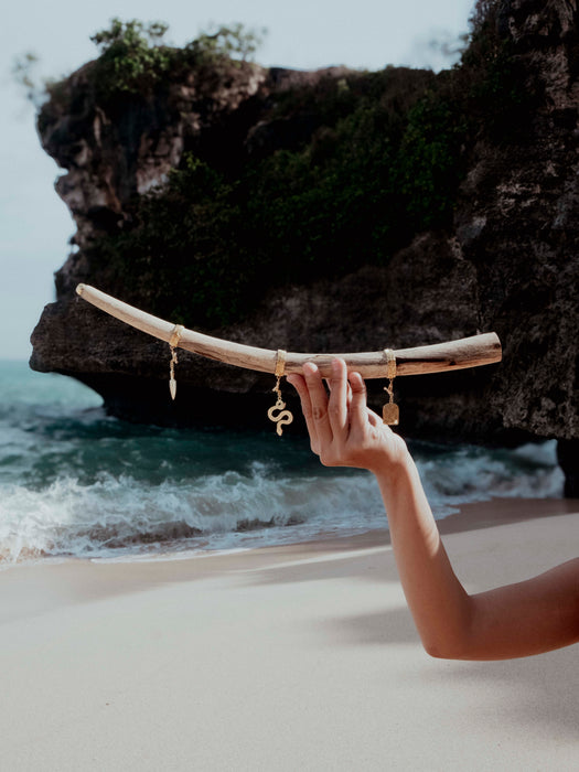 Hand holding a curved piece of driftwood with charms against a rocky beach backdrop