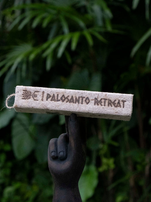 Candle labeled 'Palosanto Retreat' held by a dark hand against a blurred green leafy background