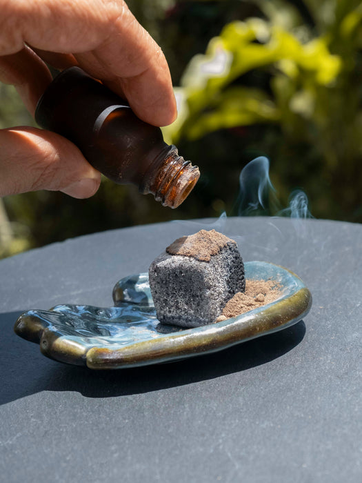 Hand pouring a liquid from a bottle onto a smoking cube on a small dish outdoors.