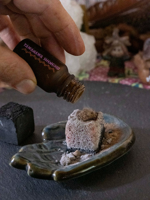 Hand pouring a liquid from a small bottle labeled 'Yawanawa Shungite' onto a stone in a decorative setting.