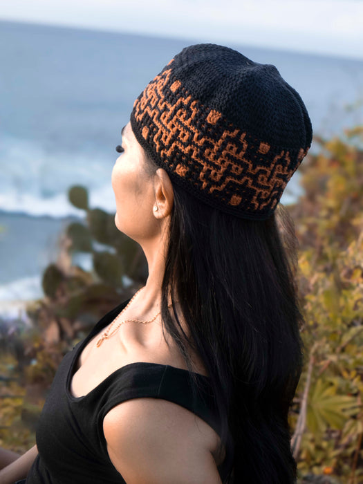 Woman wearing a black knit beanie with orange patterns, standing outdoors near water.