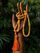 Wooden hand holding a yellow beaded mala with an orange tassel against a green leafy background