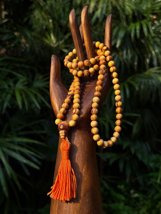 Wooden hand holding a yellow beaded mala with an orange tassel against a green leafy background