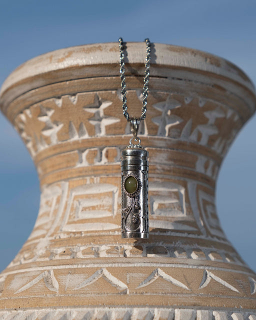 Silver pendant with a green stone on a chain against an ornate beige vase.