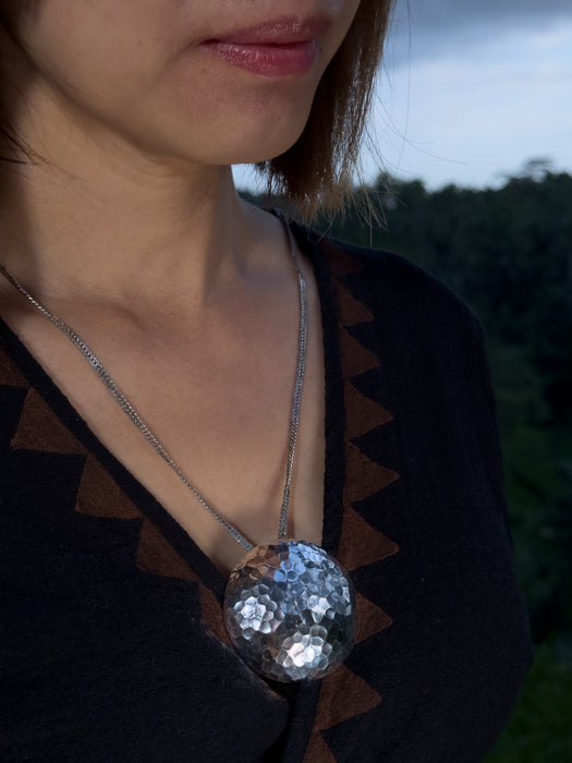 Close-up of a person wearing a necklace with a large, round, silver pendant against a blurred natural background.