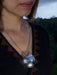 Close-up of a person wearing a necklace with a large, round, silver pendant against a blurred natural background.
