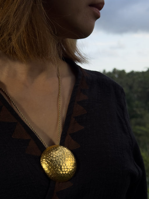 Person wearing a gold necklace with a large spherical pendant against a blurred natural background