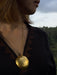 Person wearing a gold necklace with a large spherical pendant against a blurred natural background
