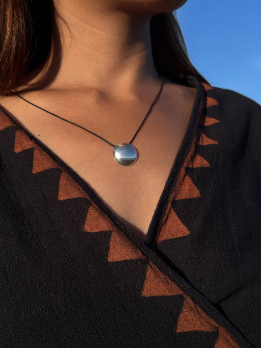 Close-up of a necklace with a circular pendant on a person wearing a black and brown patterned garment against a blue sky.