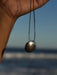 Hand holding a necklace with a silver pendant against a blurred ocean background
