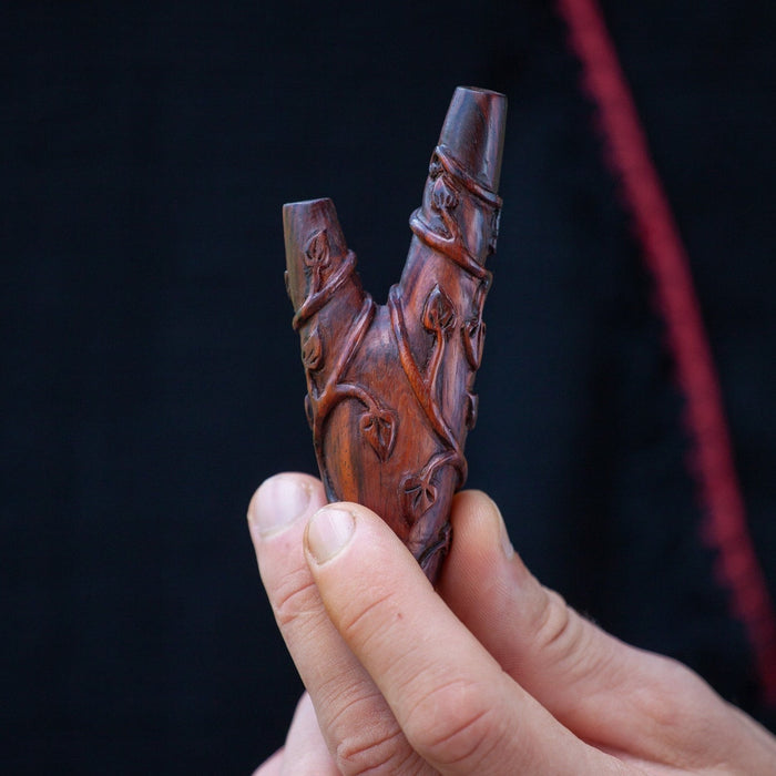 A handcrafted wooden kuripe pipe, with intricate shamanic designs, is held against a blurred green background. This unique gift, also used as a hape snuff applicator, is part of a larger collection of herbal medicine smoking accessories