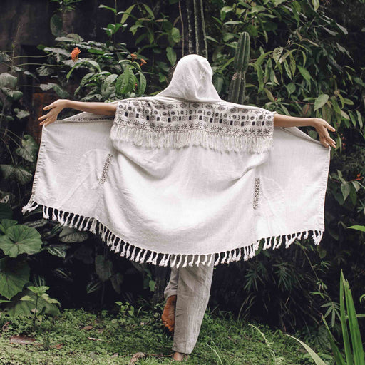 A woman stands with arms raised in a bohemian embrace, wearing a unisex linen kimono poncho with hand-embroidered tribal patterns, amidst a tranquil garden setting