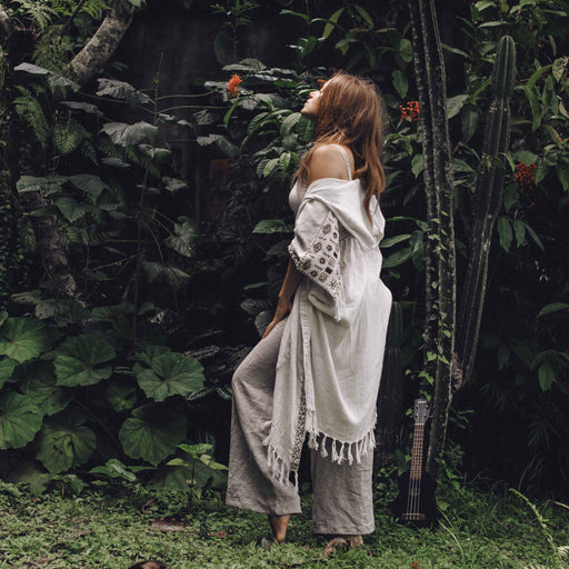 A woman stands with arms raised in a bohemian embrace, wearing a unisex linen kimono poncho with hand-embroidered tribal patterns, amidst a tranquil garden setting