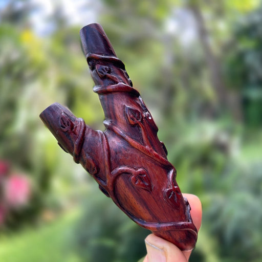 A handcrafted wooden kuripe pipe, with intricate shamanic designs, is held against a blurred green background. This unique gift, also used as a hape snuff applicator, is part of a larger collection of herbal medicine smoking accessories