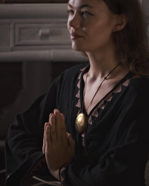 Woman in a dark outfit with a gold necklace, hands clasped together in a dimly lit room.
