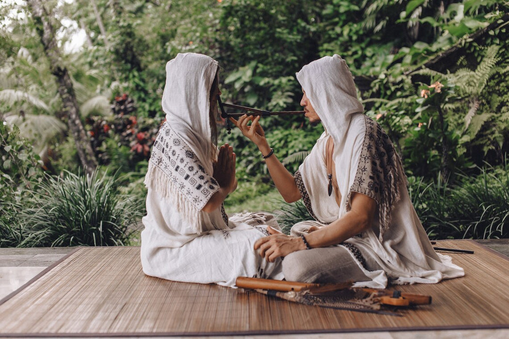Two women in white traditional attire sitting outdoors with a forest background