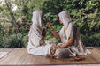 Two women in white traditional attire sitting outdoors with a forest background