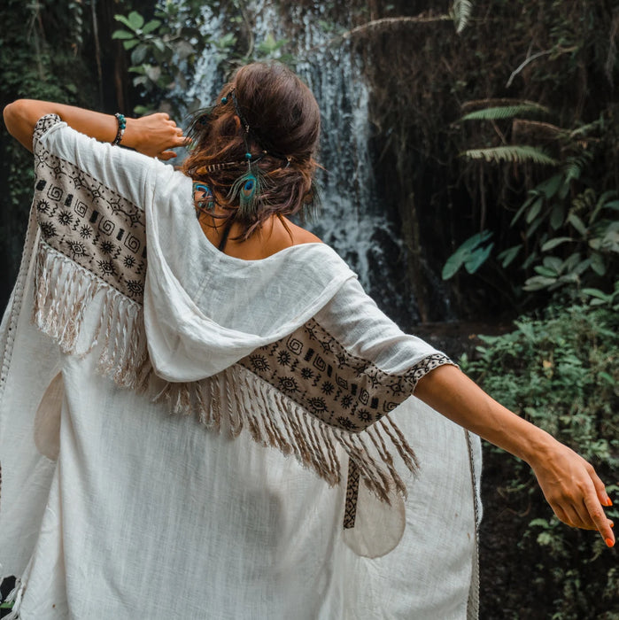 Woman in a white dress with a patterned shawl standing in front of a waterfall.