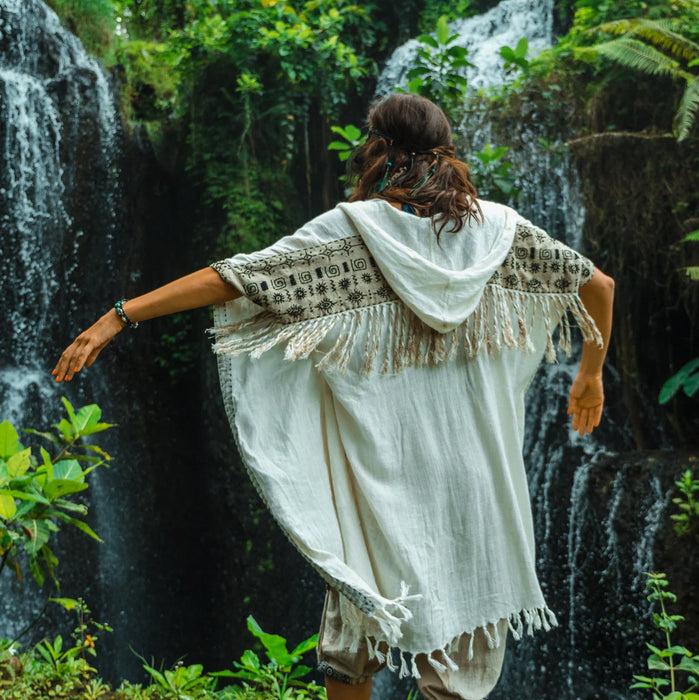 Person in a white dress with fringe standing in front of a waterfall in a lush green forest.