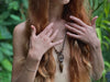 Person with long red hair and a necklace, hands covering face against a blurred green background