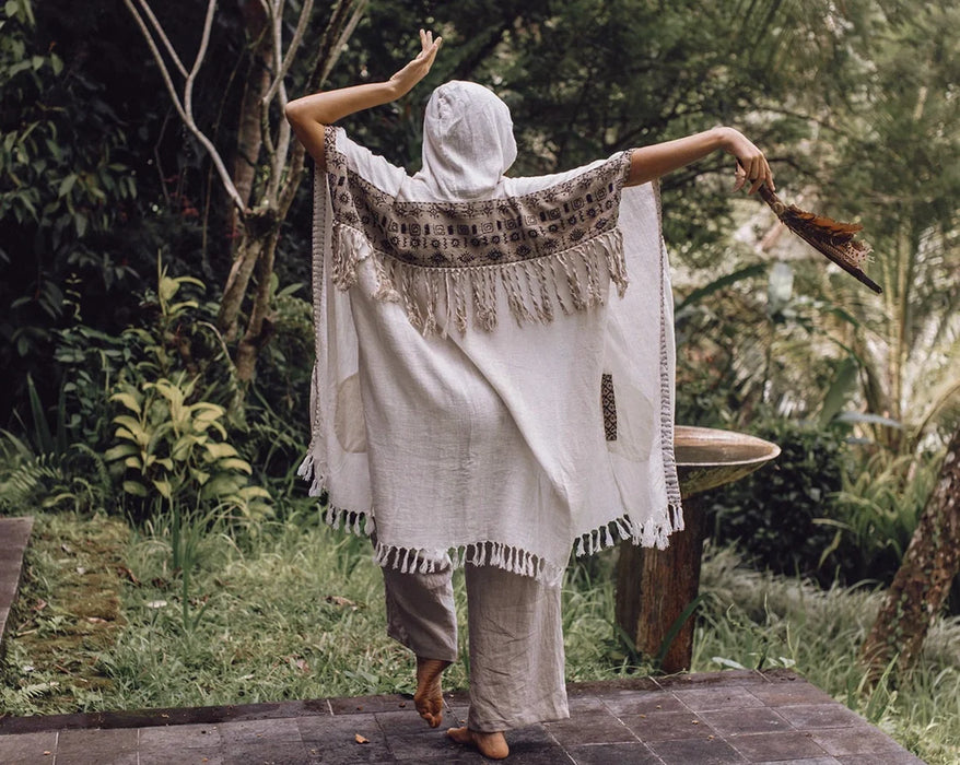 Person draped in a white shawl with fringe, standing in a natural setting.