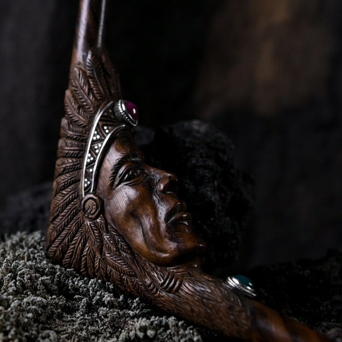 Wooden carving of a face with intricate details and a feathered headdress on a dark background