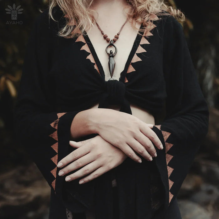 Person wearing a black top with geometric patterns and a necklace, standing in a natural setting.