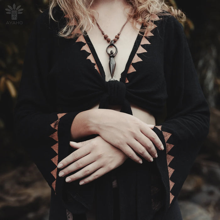 Person wearing a black top with geometric patterns and a necklace, standing in a natural setting.