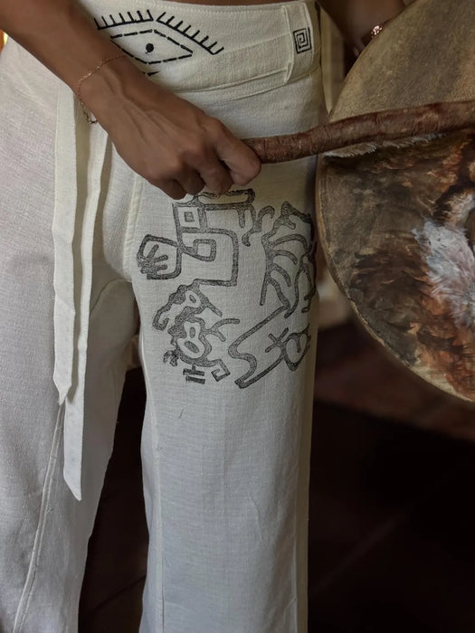 Person wearing white pants with black abstract designs, holding a wooden object.