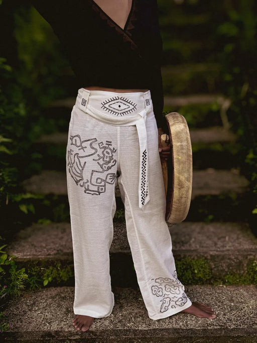 Person wearing white pants with tribal designs and a belt, holding a gong outdoors.
