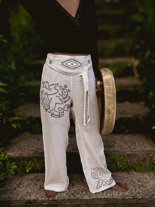 Person wearing white pants with tribal designs and a belt, holding a gong outdoors.