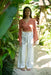 Woman in a rust-colored top and white pants with a pattern, standing in a lush green outdoor setting.